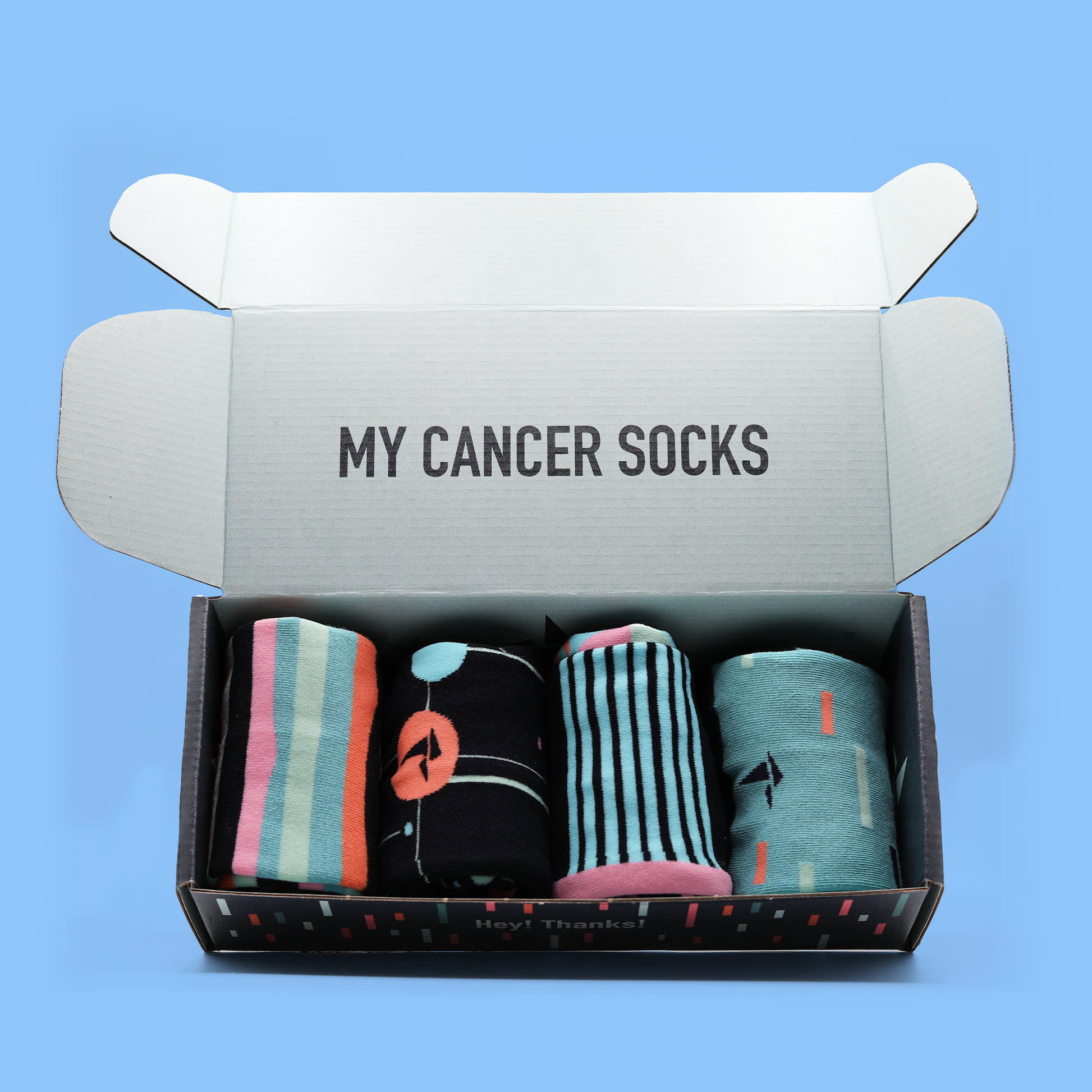 An open box of Hank's Cancer Socks in front of a blue background. The inside of the box reads "My Cancer Socks". The socks lay in a single row of four and are differing patterns with similar color schemes. The side of the box reads "Hey! Thanks!". An Awesome Socks Club product.