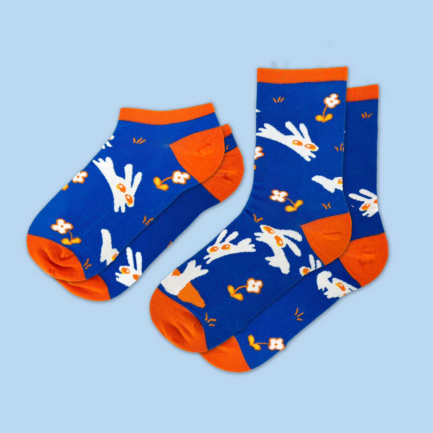 A pair of matching blue and orange socks against a light blue background. The socks feature a playful pattern of white rabbits with orange ears and noses, along with small orange flowers and decorative elements scattered across the bright blue fabric. The socks have contrasting orange heels, toes, and cuffs. One sock is positioned as an ankle sock style while the other is a crew sock length, both displaying the same whimsical rabbit and floral design