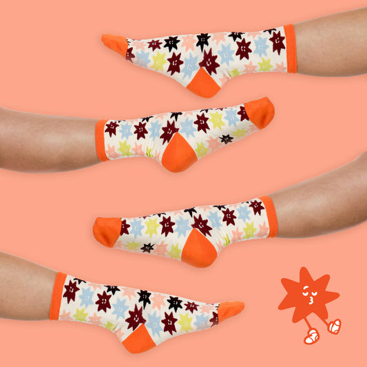 Four pairs of crew socks displayed on human feet against a peach background. The socks have a white base with a colorful pattern of cartoon suns and stars in orange, red, black, blue, and green, with solid orange heels, toes, and cuffs.