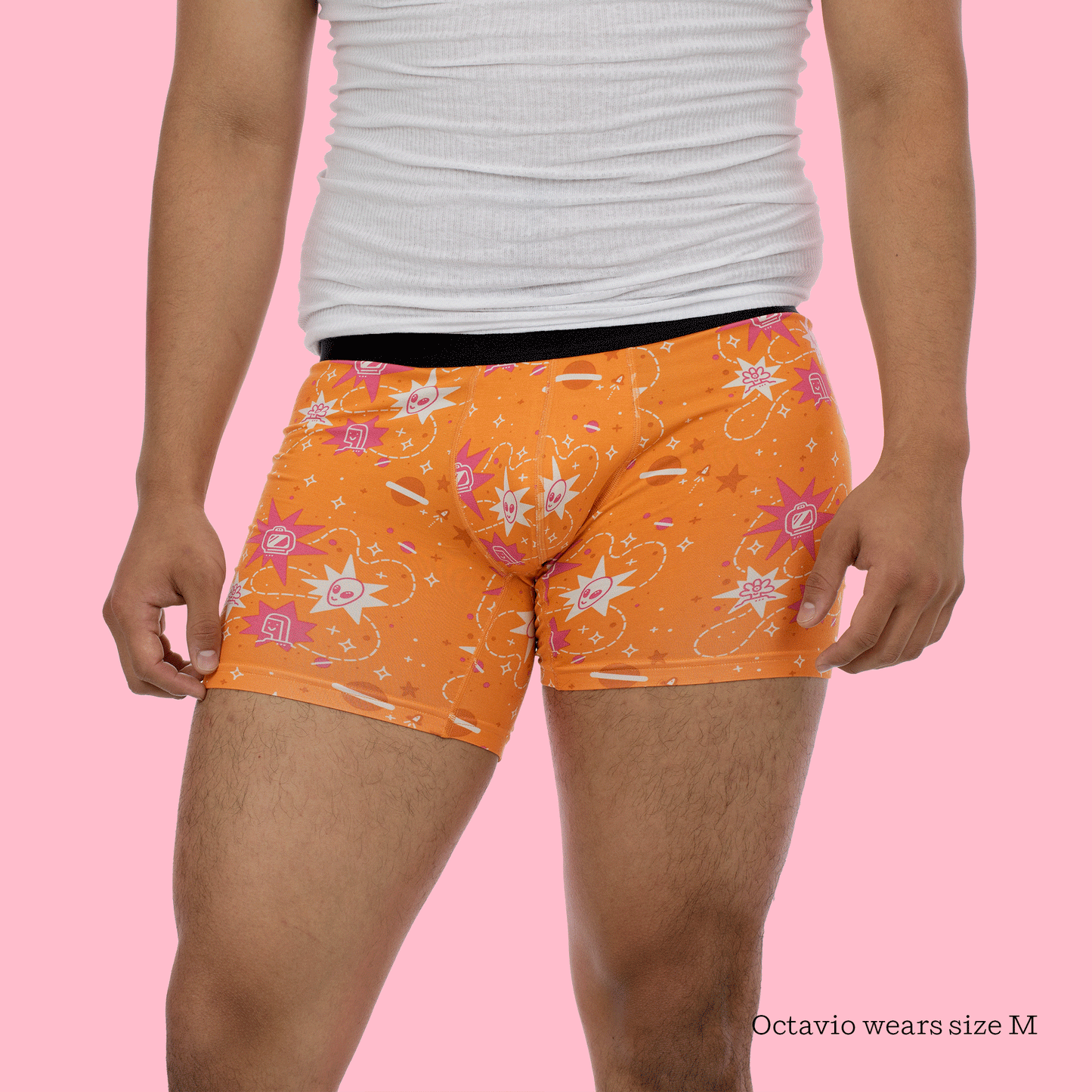 A person wearing a white tank top and orange boxer shorts with the same star and symbol pattern, photographed against a pink background. The model has their hands positioned at their hips. Text indicates "Octavio wears size M.