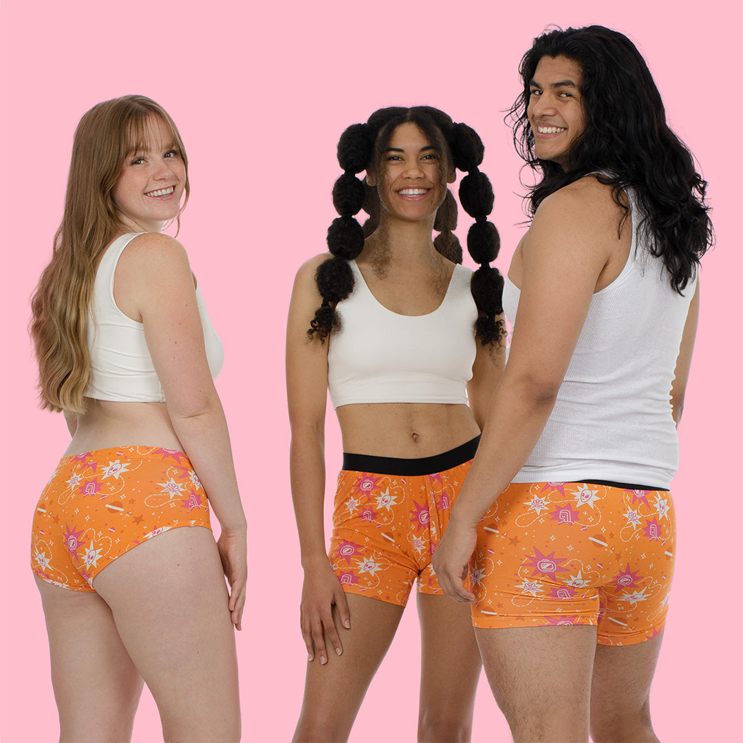 he same three people, now photographed from the front, all smiling at the camera. They're wearing the same orange patterned underwear/shorts with white tops against the pink background, showing the inclusive sizing and diverse representation of the garment line.