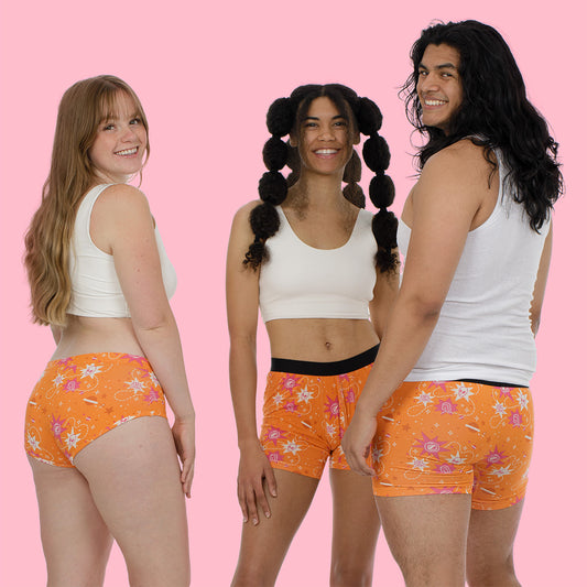 he same three people, now photographed from the front, all smiling at the camera. They're wearing the same orange patterned underwear/shorts with white tops against the pink background, showing the inclusive sizing and diverse representation of the garment line.