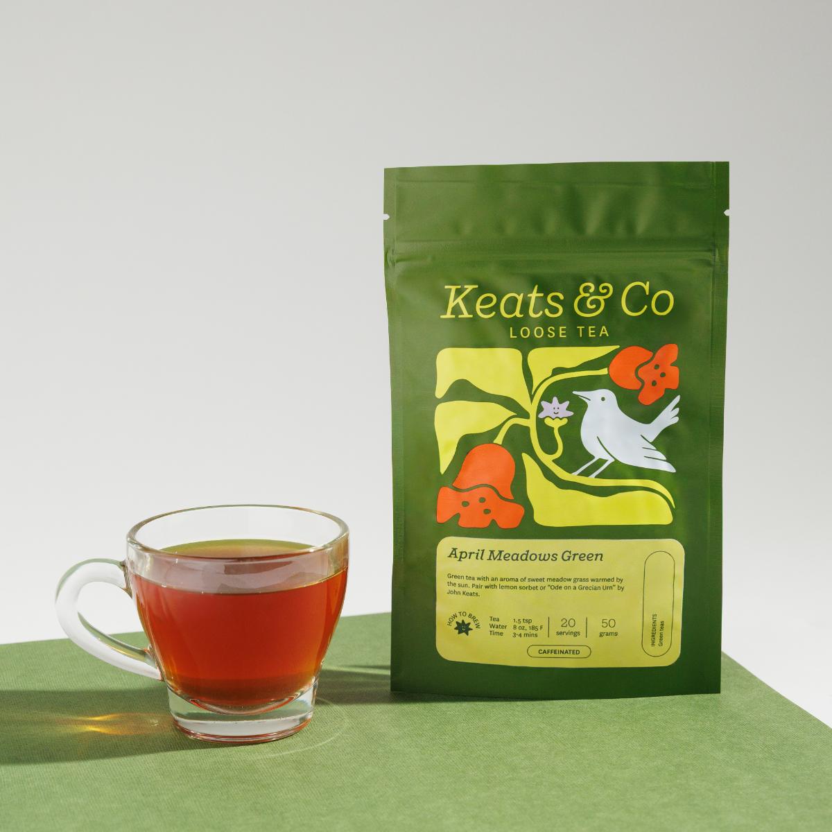 Green pouch of Keats & Co Loose Tea April Meadows Green against a white background, featuring illustrated artwork of a white bird and orange flowers, displayed next to a clear glass mug filled with redish brewed tea on a green surface.