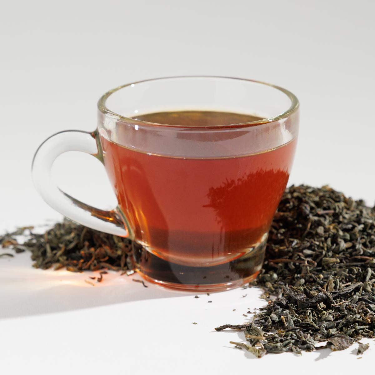 Glass cup of redish April Meadows Green tea positioned next to a pile of loose leaf tea blend on a reflective white surface against a soft white background. A Good Store product.