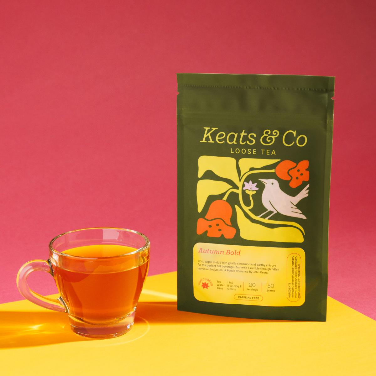 Green pouch of Keats & Co Loose Tea Autumn Bold against a pink background, featuring illustrated artwork of a white bird and orange flowers, displayed next to a clear glass mug filled with yellowish brewed tea on a yellow surface.