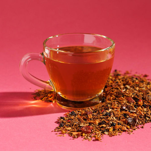 Glass cup of orange-ish Autumn Bold tea positioned next to a pile of loose herbals on a pink bacground. A Good Store product.