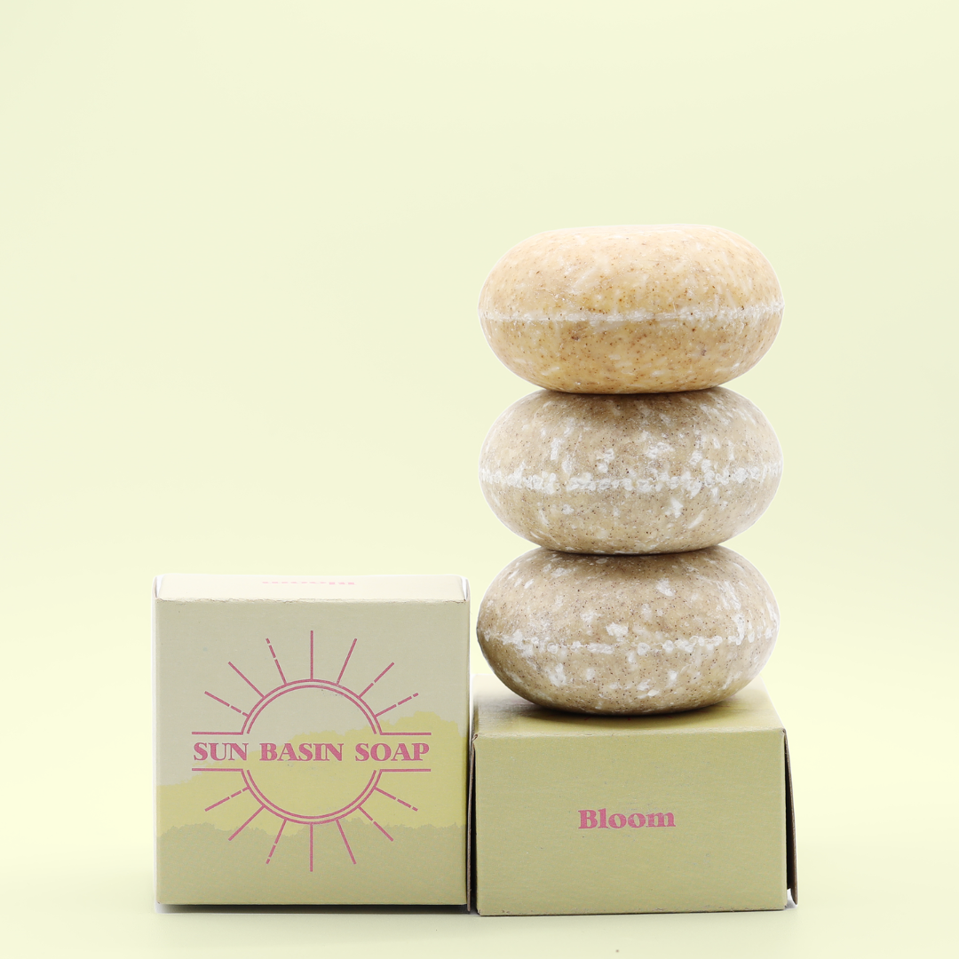 A stack of three of the Bloom Shampoo Bars on top of a small, yellow box that reads Bloom. There is a small box next to it with the Sun Basin Soap logo in pink. A Good Store product. 