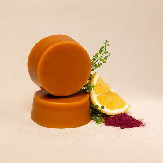 Two round orange-brown conditioner bars stacked on a beige surface, styled with a lemon slice, green sprigs, and a small pile of dark magenta powder against a plain beige background.
