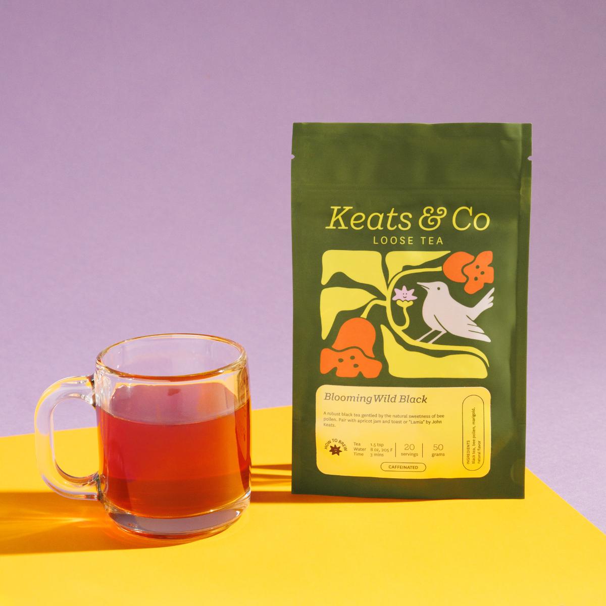 Green pouch of Keats & Co Loose Tea Blooming Wild Black against a purple background, featuring illustrated artwork of a white bird and orange flowers, displayed next to a clear glass mug filled with red-ish brewed tea on a yellow surface.