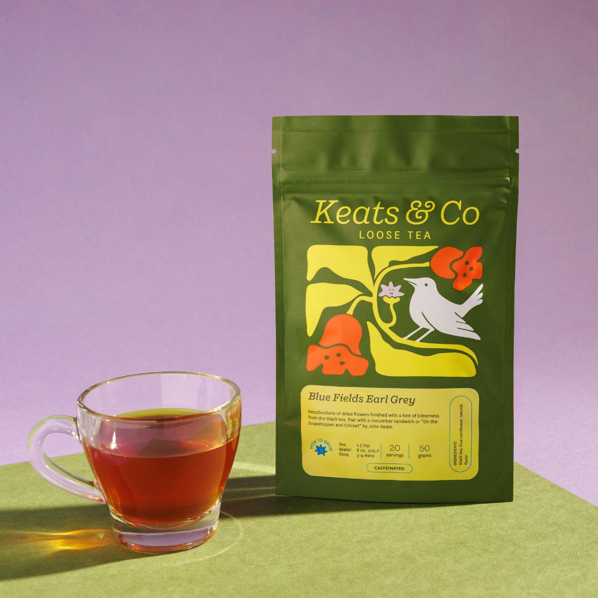 Green pouch of Keats & Co Loose Tea Blue Fields Early Grey against a purple background, featuring illustrated artwork of a white bird and orange flowers, displayed next to a clear glass mug filled with amberish brewed tea on a green surface.