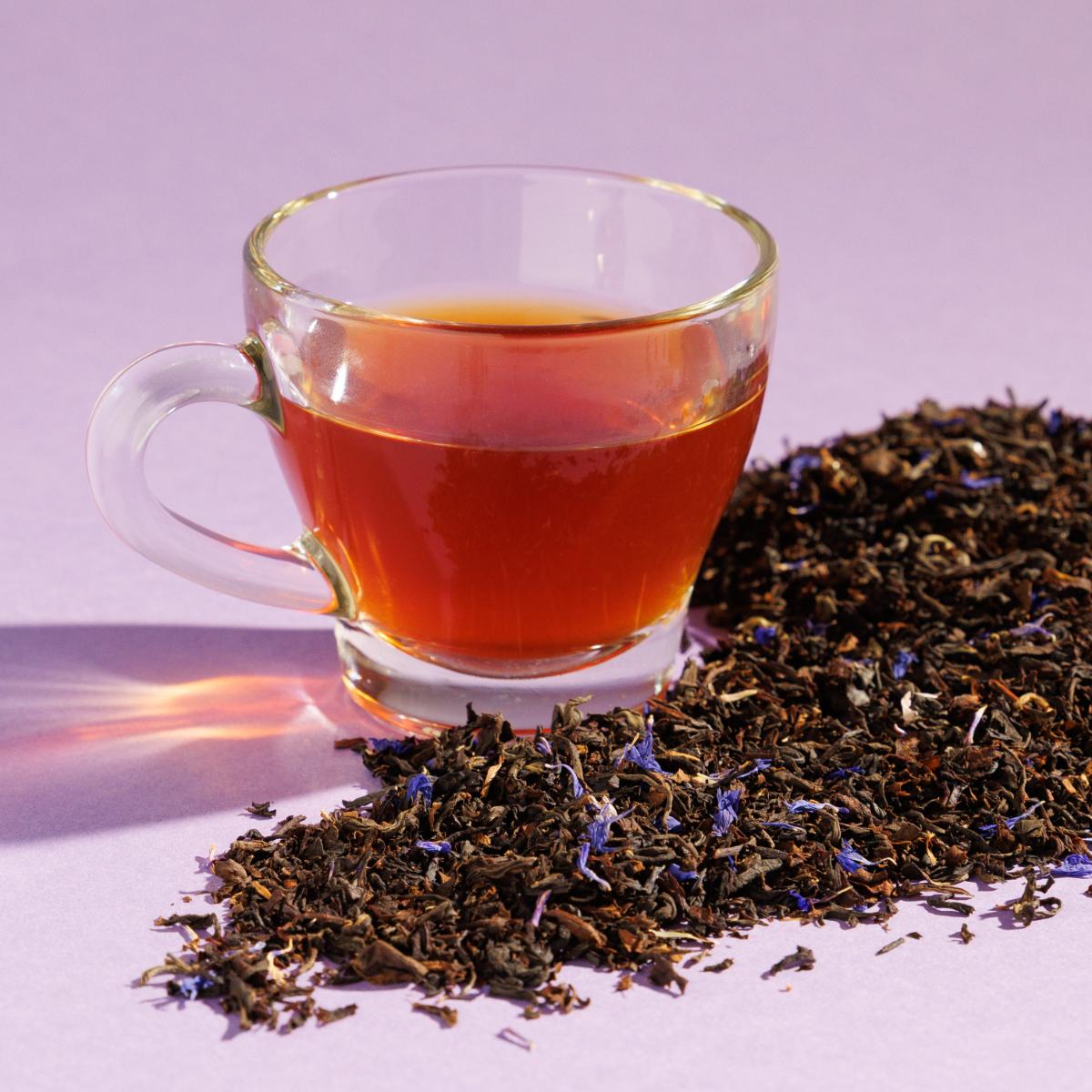 Glass cup of amber Blue Fields Earl Grey tea positioned next to a pile of loose leaf tea blend containing specks of purple on a purple background. A Good Store product.