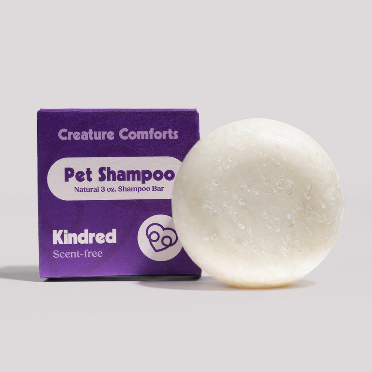 A round, off-white natural shampoo bar sits next to its purple packaging box. The box is labeled 'Creature Comforts Pet Shampoo' and features the brand 'Kindred'.