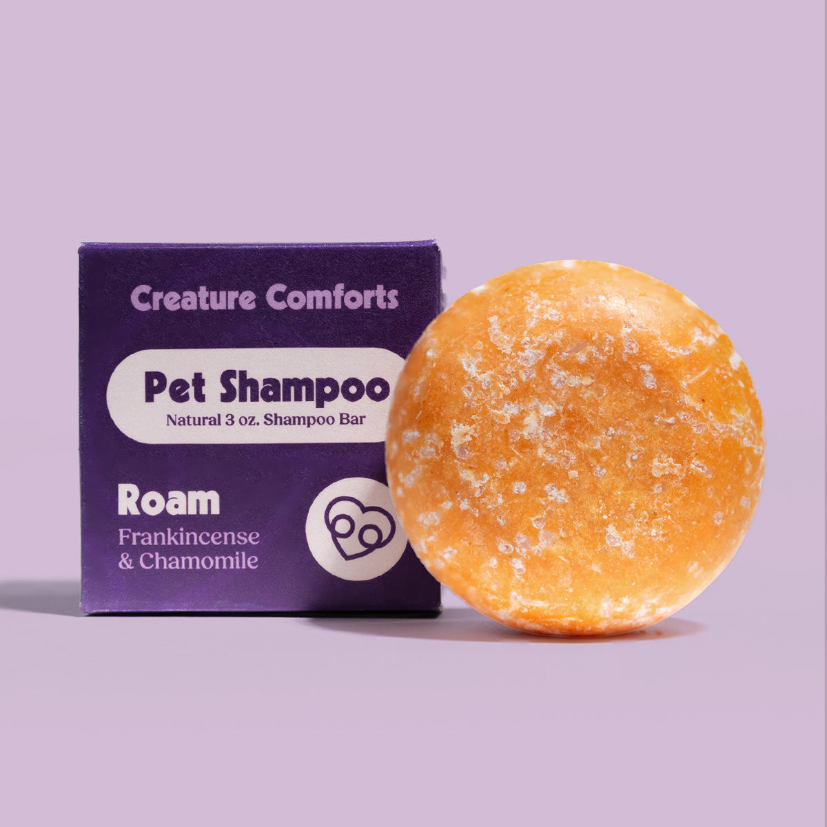 A round orange pet shampoo bar with stands next to its purple packaging box labeled 'Creature Comforts Pet Shampoo Roam Frankincense & Chamomile' on a solid lavender background.