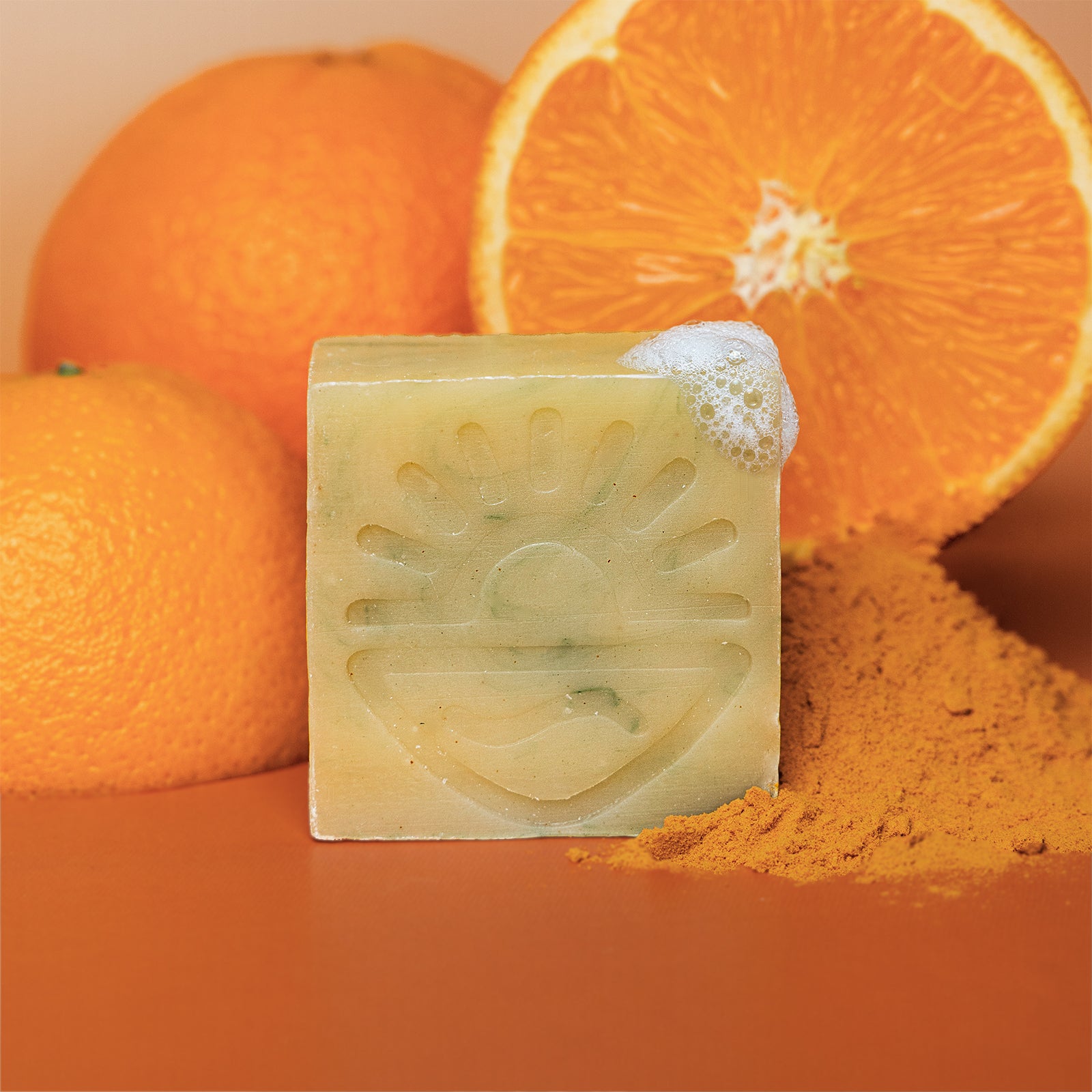 A rectangular bar of pale yellow soap with an embossed sun and wave design, shown with orange slices, whole oranges, and orange powder on an orange background.