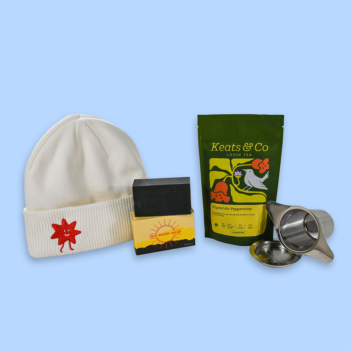 Cream beanie with red logo, soap bar, green tea package, and metal strainer on a light blue background