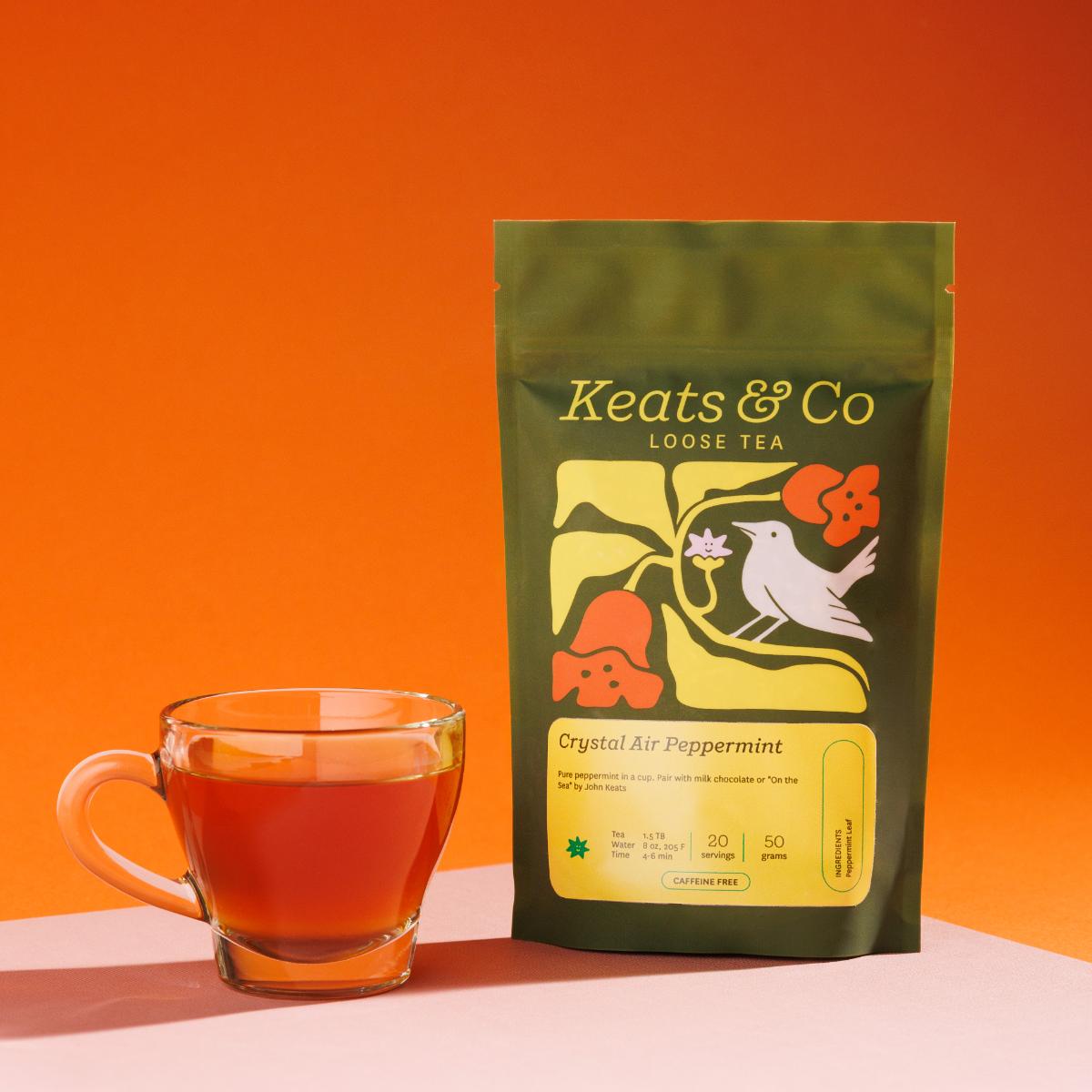 Green pouch of Keats & Co Loose Tea Crystal Air Peppermint against an orange background, featuring illustrated artwork of a white bird and orange flowers, displayed next to a clear glass mug filled with an orange brewed tea on a pink surface.