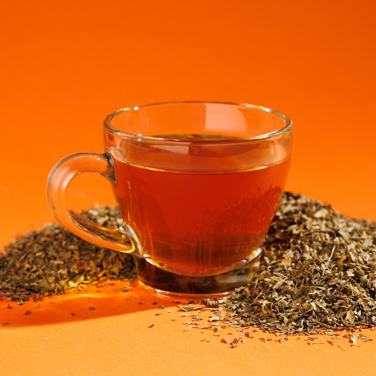 Glass cup of redish Crystal Air Peppermint tea positioned next to a pile of loose leaf tea blend containing peppermint leaves on an orange surface against an orange background. A Good Store product.