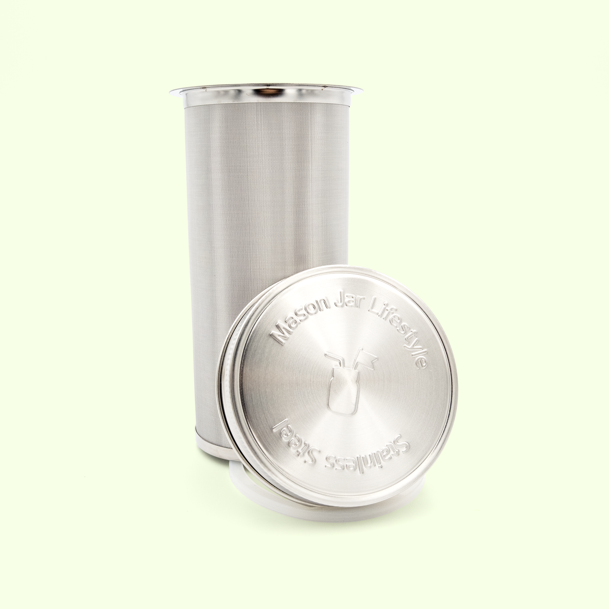 A stainless steel infuser with fine mesh sides and a solid lid, positioned next to its matching lid on a pale yellow background. The lid is embossed with "Mason Jar Lifestyle" text and appears to fit standard mason jars.