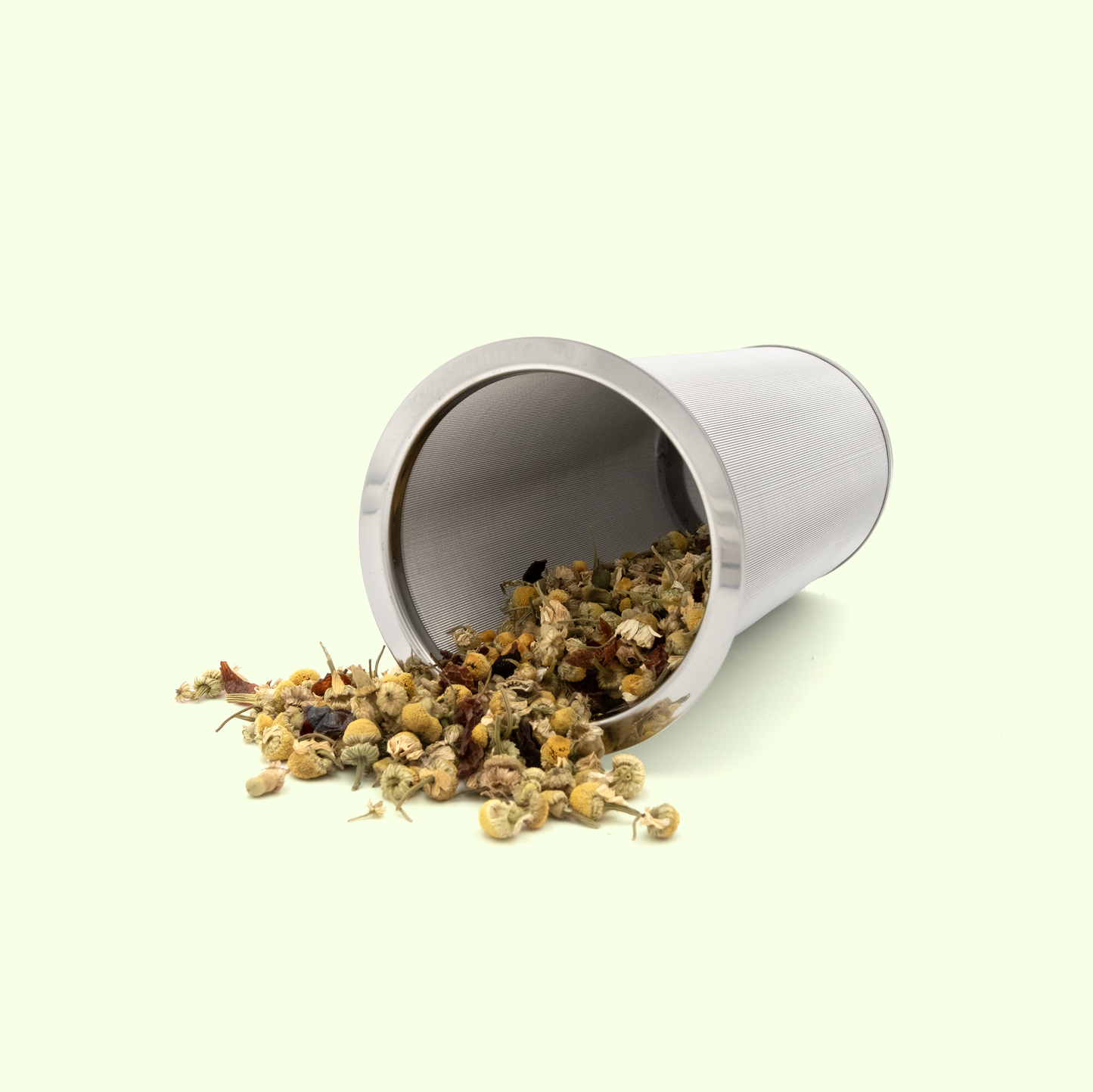 A cylindrical stainless steel tea infuser tipped on its side, spilling dried chamomile flowers and herbs onto a pale yellow background. The mesh strainer contains a mixture of small yellow chamomile buds and other herbal tea ingredients.