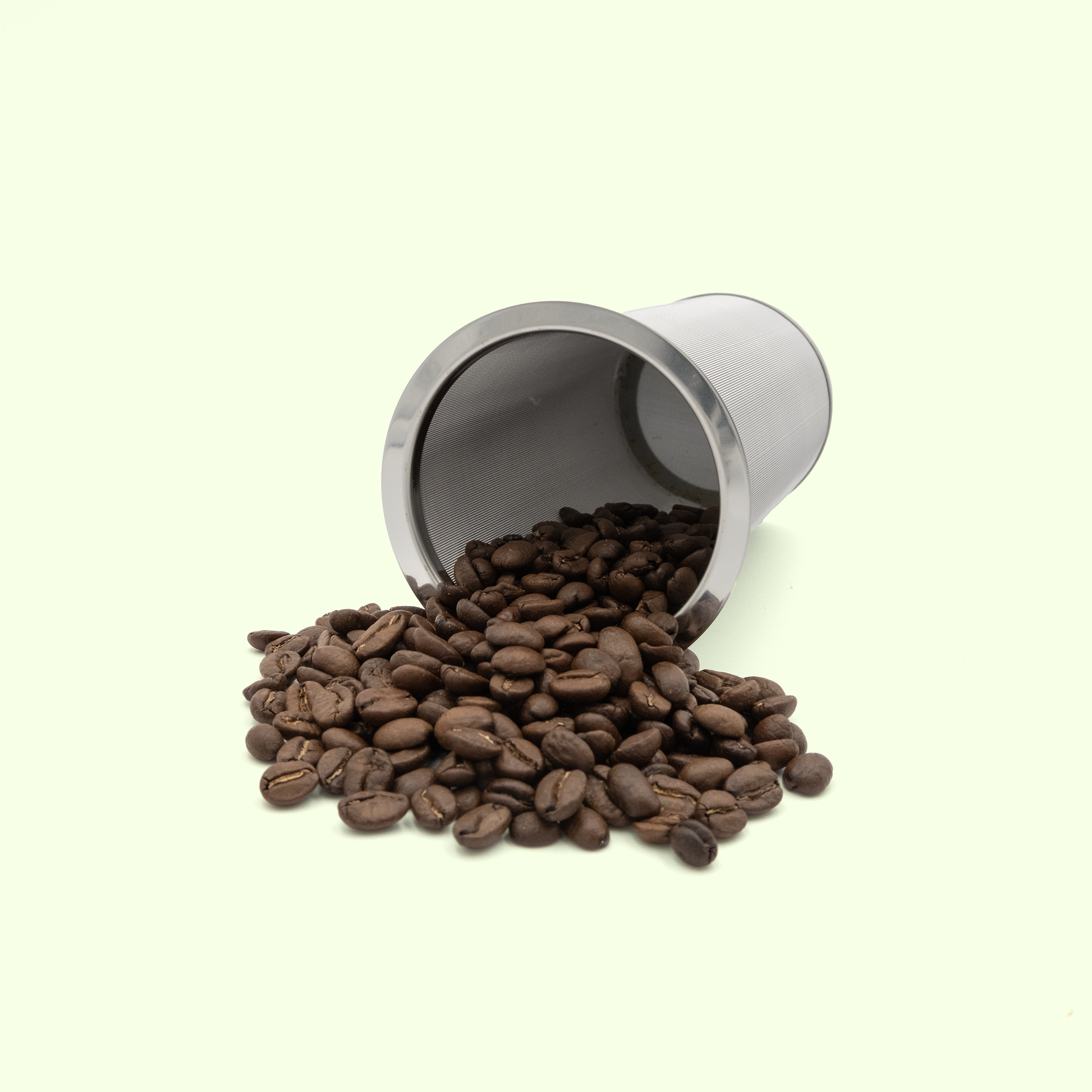 A stainless steel infuser tipped over, this time spilling dark roasted coffee beans onto a pale yellow background. The mesh cylinder shows its fine filtering capability for both loose leaf teas and coffee grounds.