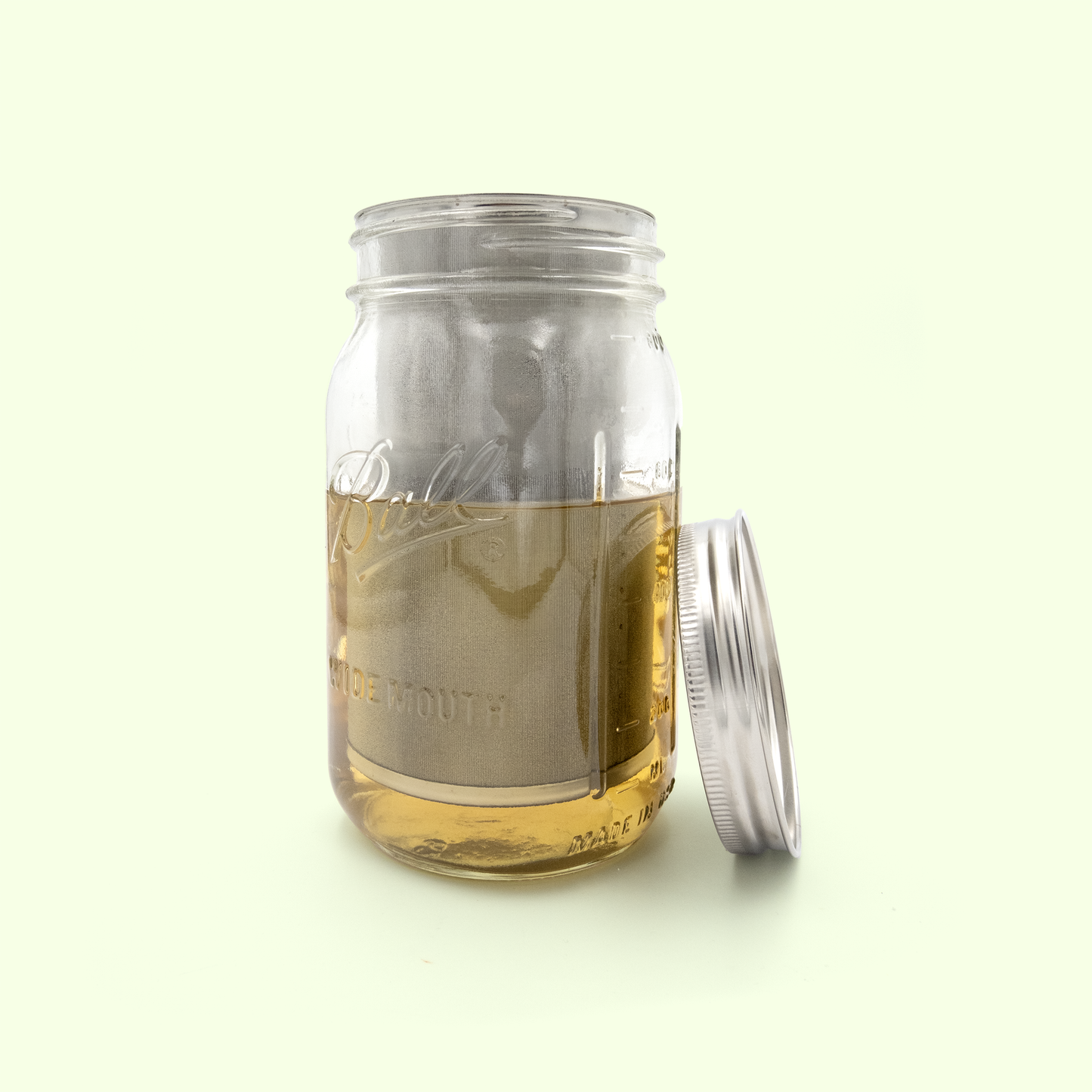 A clear glass mason jar containing golden-colored tea or herbal infusion, with the stainless steel mesh tea infuser visible inside. The mason jar's metal lid sits beside it on a pale yellow background.