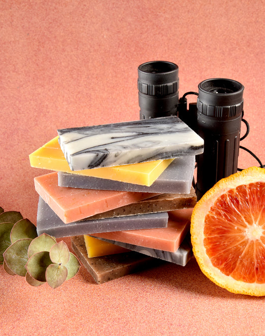 A stack of all of the different Sun Basin Soap scents in thin bars for the sample pack. A pair of binoculars, a slice of blood orange and a eucalyptus are surrounding the soaps. They all sit in front of a salmon background. A Good Store subscription product.