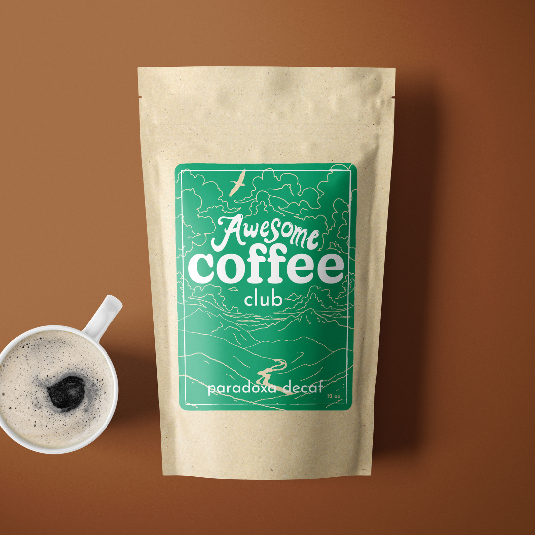 A photo of a brown bag of coffee with a green label that shows a mountain scene and has the text "Awesome Coffee Club; Paradoxa Decaf". The bag sites atop a rust colored background with a cup of coffee next to it. A Good Store subscription product.