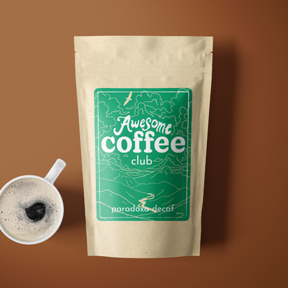 A photo of a brown bag of coffee with a green label that shows a mountain scene and has the text "Awesome Coffee Club; Paradoxa Decaf". The bag sites atop a rust colored background with a cup of coffee next to it. A Good Store subscription product.