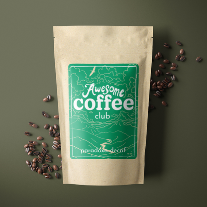 A photo of a brown bag of coffee with a green label that shows a mountain scene and has the text "Awesome Coffee Club; Paradoxa Decaf". The bag sites atop a dark green background with whole coffee beans spread around it. A Good Store subscription product.