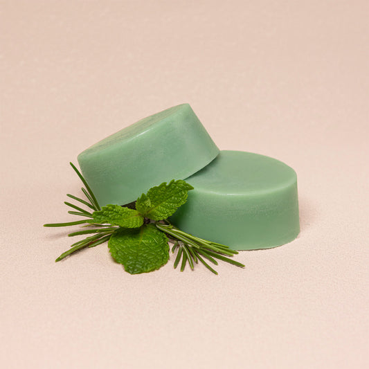 Two mint green conditioner bars stacked with fresh green mint leaves and rosemary sprigs, set against a solid pale pink background.