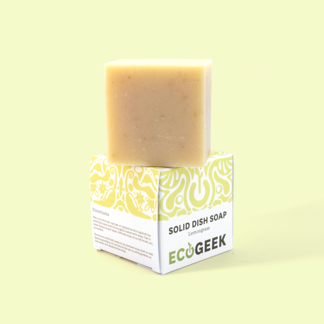 The image shows a bar of Lemongrass solid dish soap from the brand "ECOGEEK." The soap is a light beige/cream color and sits atop its green and white packaging. The packaging features a decorative green swirly pattern on the top portion and "SOLID DISH SOAP [Lemongrass]" written prominently. The ECOGEEK logo appears at the bottom of the packaging. The entire product is photographed against a pale yellow background, creating a clean, minimalist presentation. Solid dish soap from EcoGeek a Good Store product.