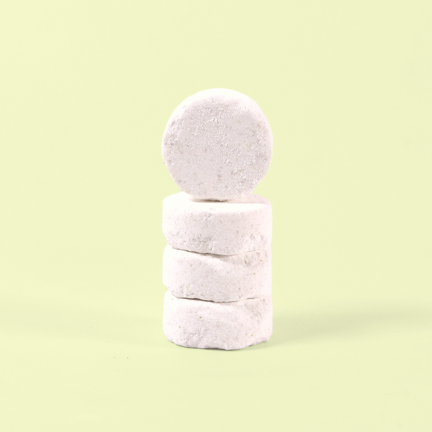 A stack of four white dishwasher tablets photographed against a pale yellow background. The circular tablets have a slightly textured, compressed powder appearance typical of eco-friendly dishwasher detergent. They're stacked vertically to show their thickness and uniform white color. Dishwasher tablets from EcoGeek a Good Store product.