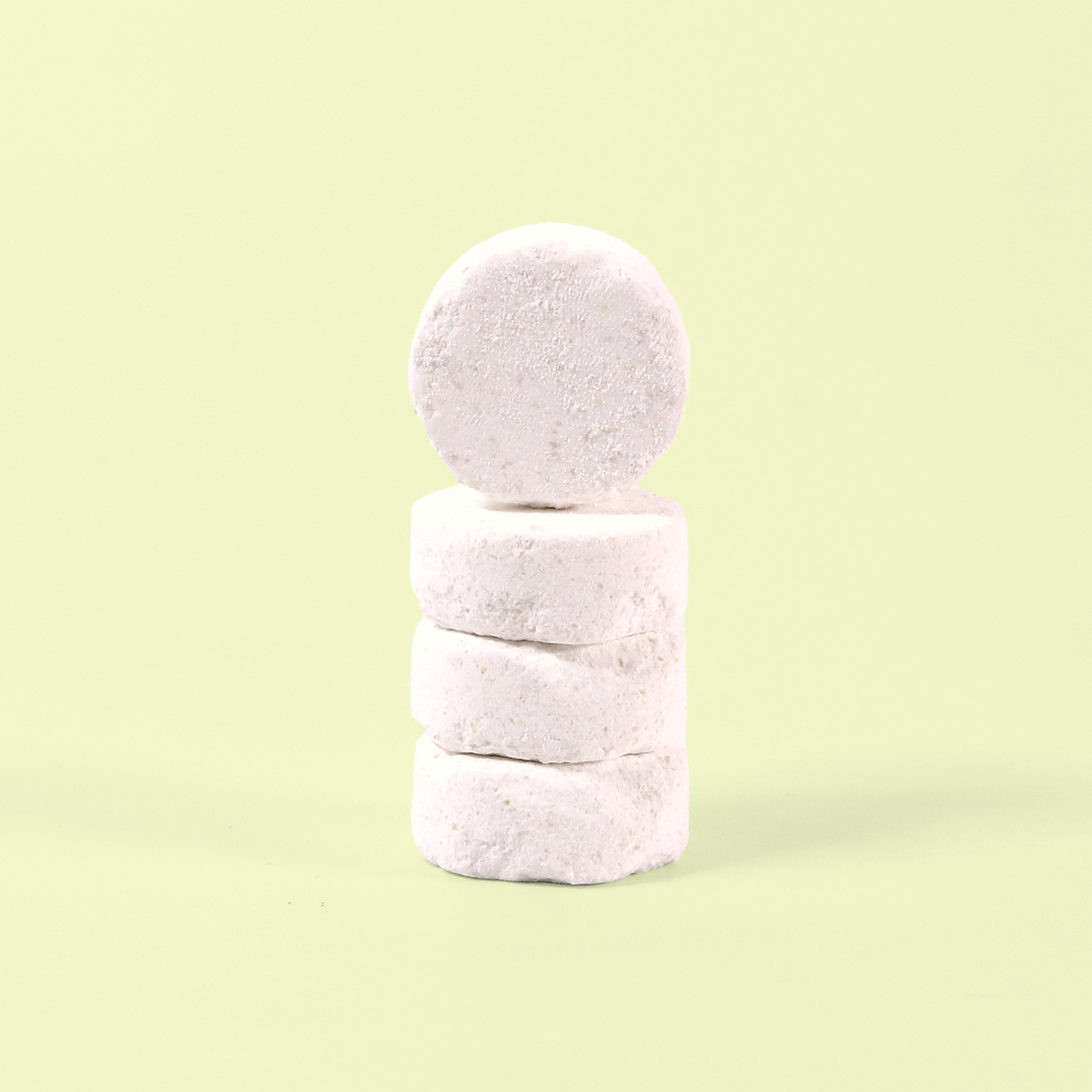 A stack of four white dishwasher tablets photographed against a pale yellow background. The circular tablets have a slightly textured, compressed powder appearance typical of eco-friendly dishwasher detergent. They're stacked vertically to show their thickness and uniform white color. Dishwasher tablets from EcoGeek a Good Store product.