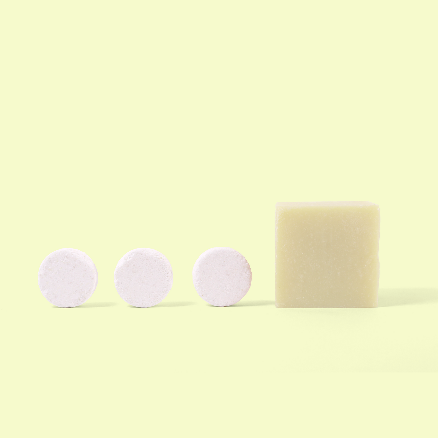 Three white circular dishwasher tablets arranged in a row next to a pale yellow rectangular soap bar against a light yellow background. The minimalist composition shows the contrast between the different eco-friendly cleaning product forms - the compressed powder tablets and the solid dish soap. Dish bundle from EcoGeek a Good Store product.