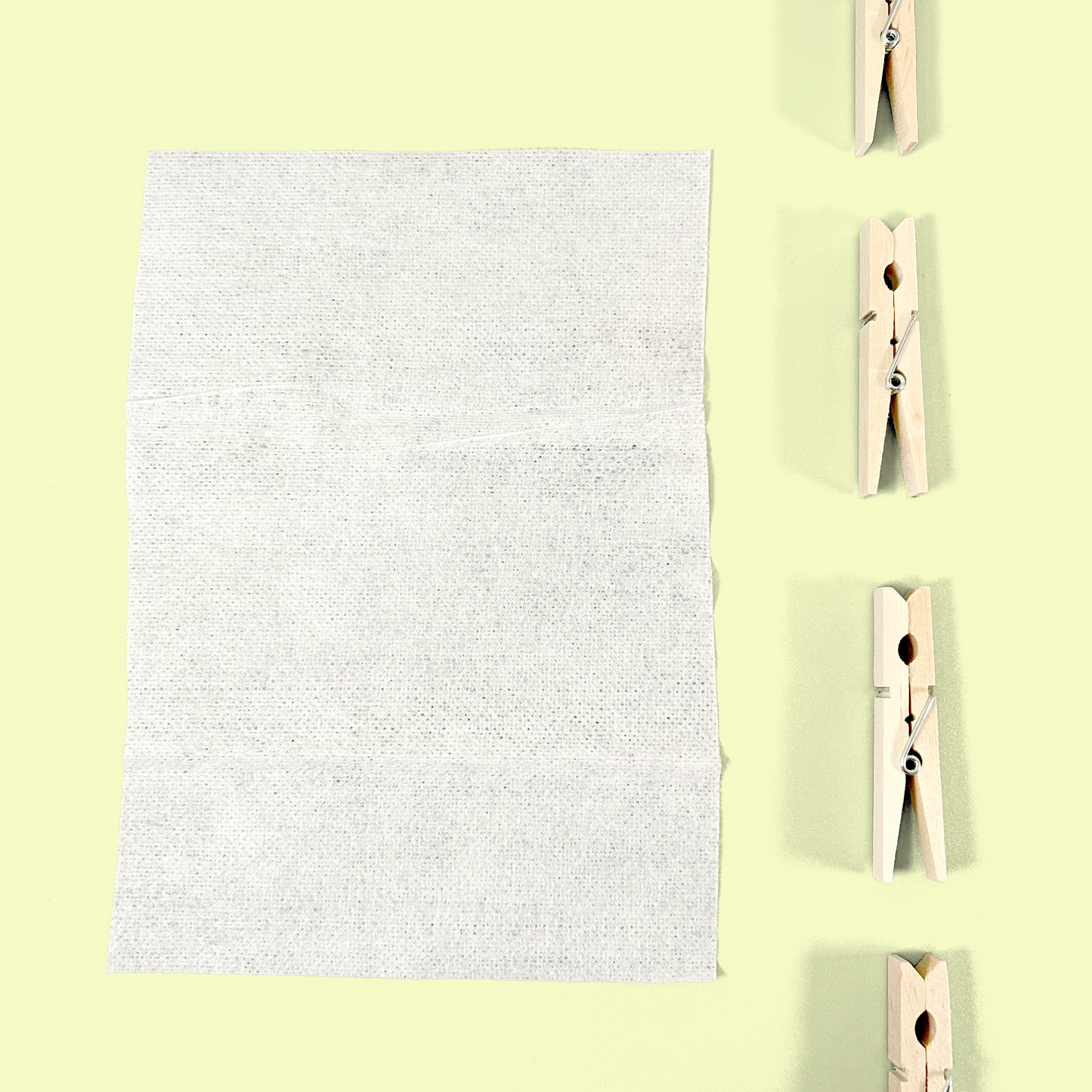 A close-up view of a single white fabric softening dryer sheet unfolded to show its full size and textured surface. Four wooden clothespins are arranged vertically to the right against a pale yellow background, showing how the product might be used on a clothesline.
Dryer sheets from EcoGeek a Good Store product.