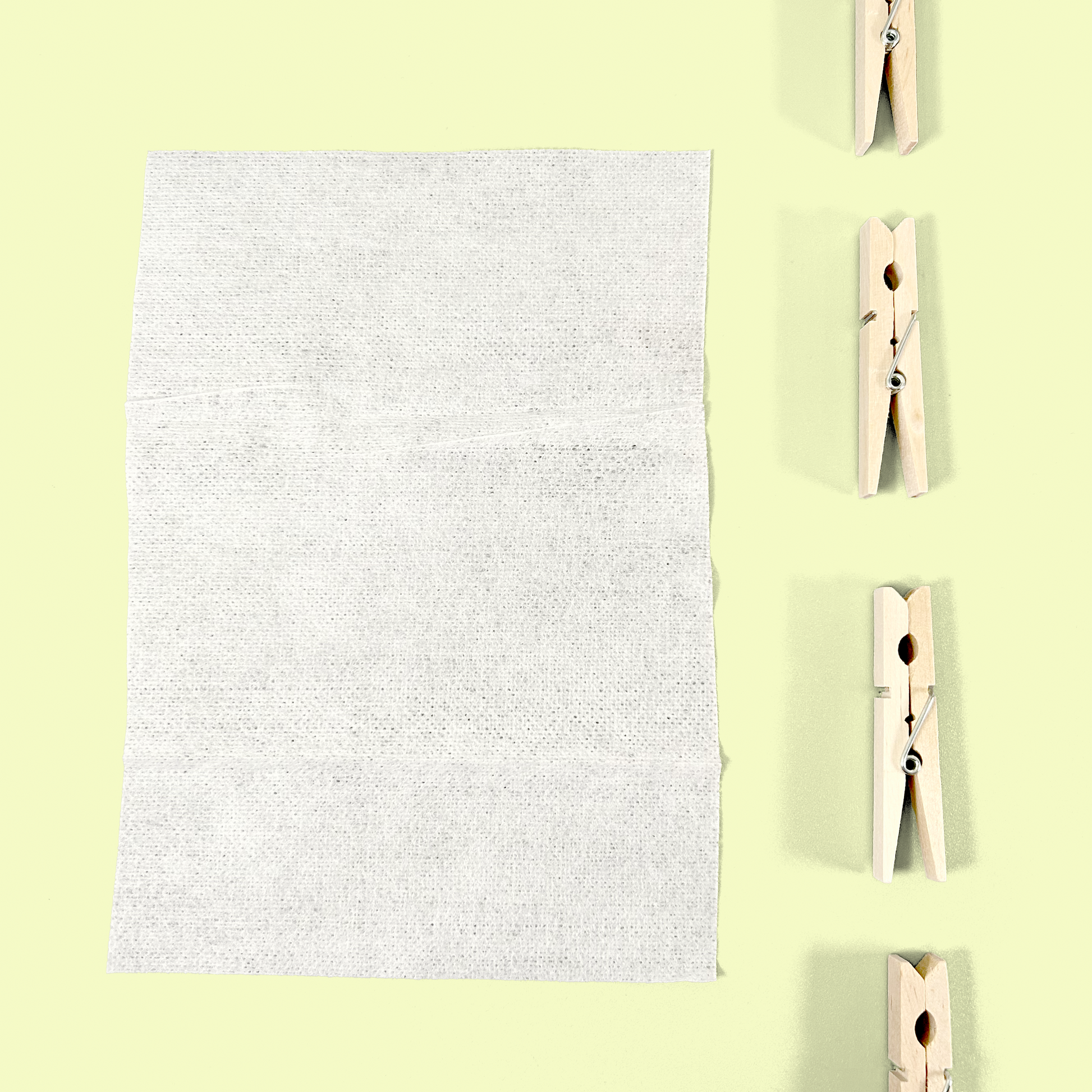 A close-up view of a single white fabric softening dryer sheet unfolded to show its full size and textured surface. Four wooden clothespins are arranged vertically to the right against a pale yellow background, showing how the product might be used on a clothesline.
Dryer sheets from EcoGeek a Good Store product.