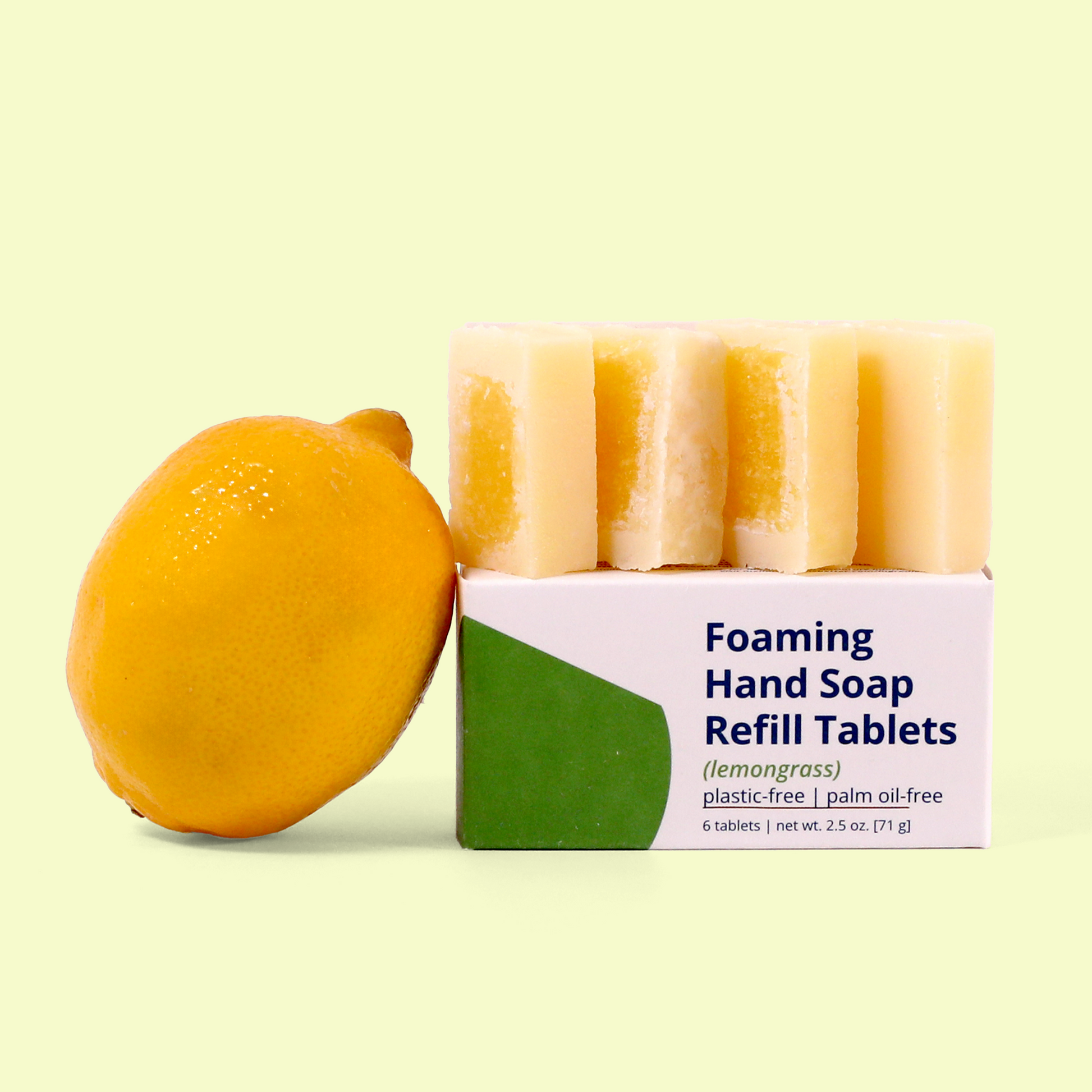 A box of Meliora lemongrass scented Foaming Hand Soap Refill Tablets with a lemon placed beside it against a pale yellow background. The white and green packaging specifies "plastic-free | palm oil-free" and "6 tablets | net wt. 2.5 oz. (71g)." Five light yellow soap tablets with darker yellow centers are displayed on top of the box. Foaming hand soap refill tablets from EcoGeek a Good Store product. 