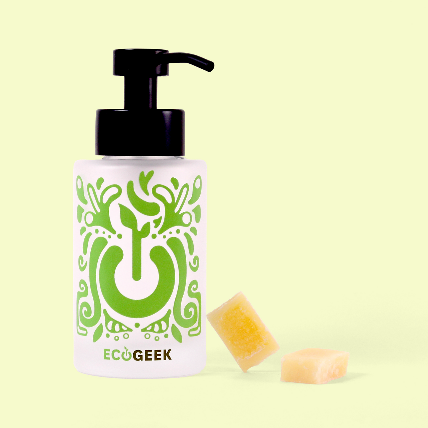 A glass pump bottle with a black dispenser top featuring a decorative green pattern with a power button symbol integrated into the design accompanied by two small beige/yellow soap concentrate cubes positioned to the right of the bottle. The entire arrangement is displayed against a pale yellow background. Foaming hand soap bottle from EcoGeek a Good Store product.