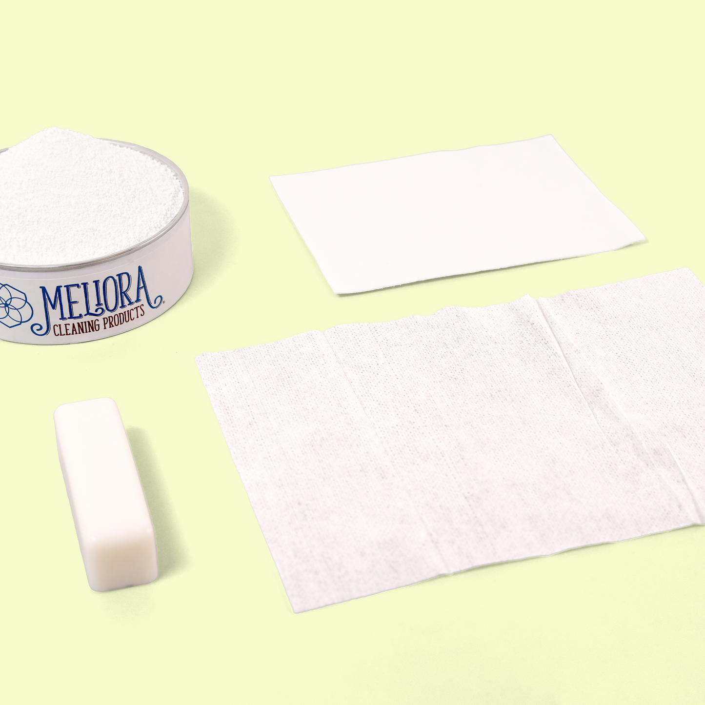 A collection of eco-friendly laundry products. The image shows a white Meliora Cleaning Products container lid filled with white powder, a rectangular white soap stick laundry stain remover, and two white fabric or paper sheets of different textures - possibly a dryer sheet and a cleaning cloth - demonstrating the company's eco-friendly cleaning solutions. Laundry bundle from EcoGeek a Good Store product.