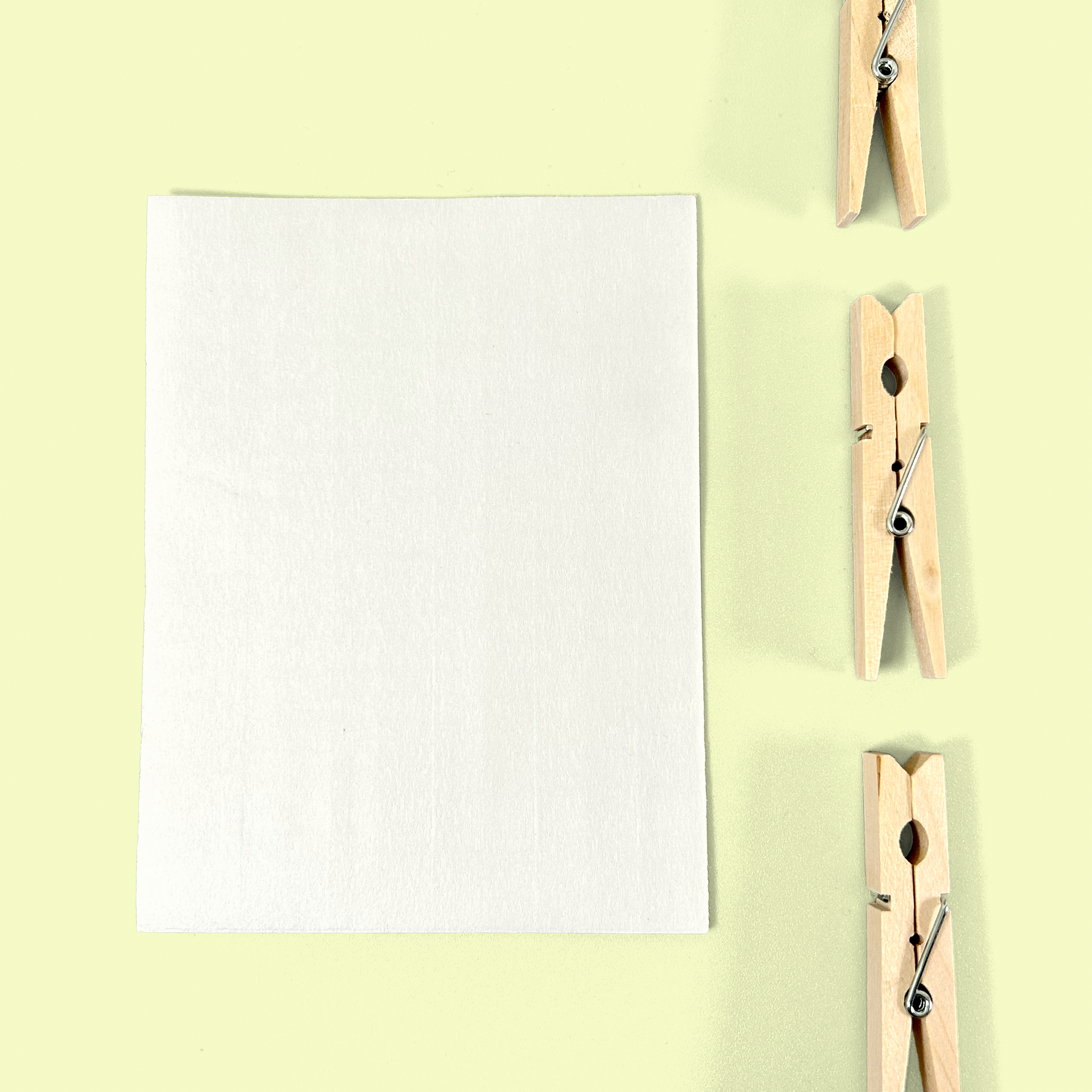 A single white laundry detergent sheet displayed flat on a pale yellow background with three wooden clothespins arranged vertically to the right. The image showcases the thin, paper-like texture of the detergent sheet. Laundry detergent sheets from EcoGeek a Good Store product.