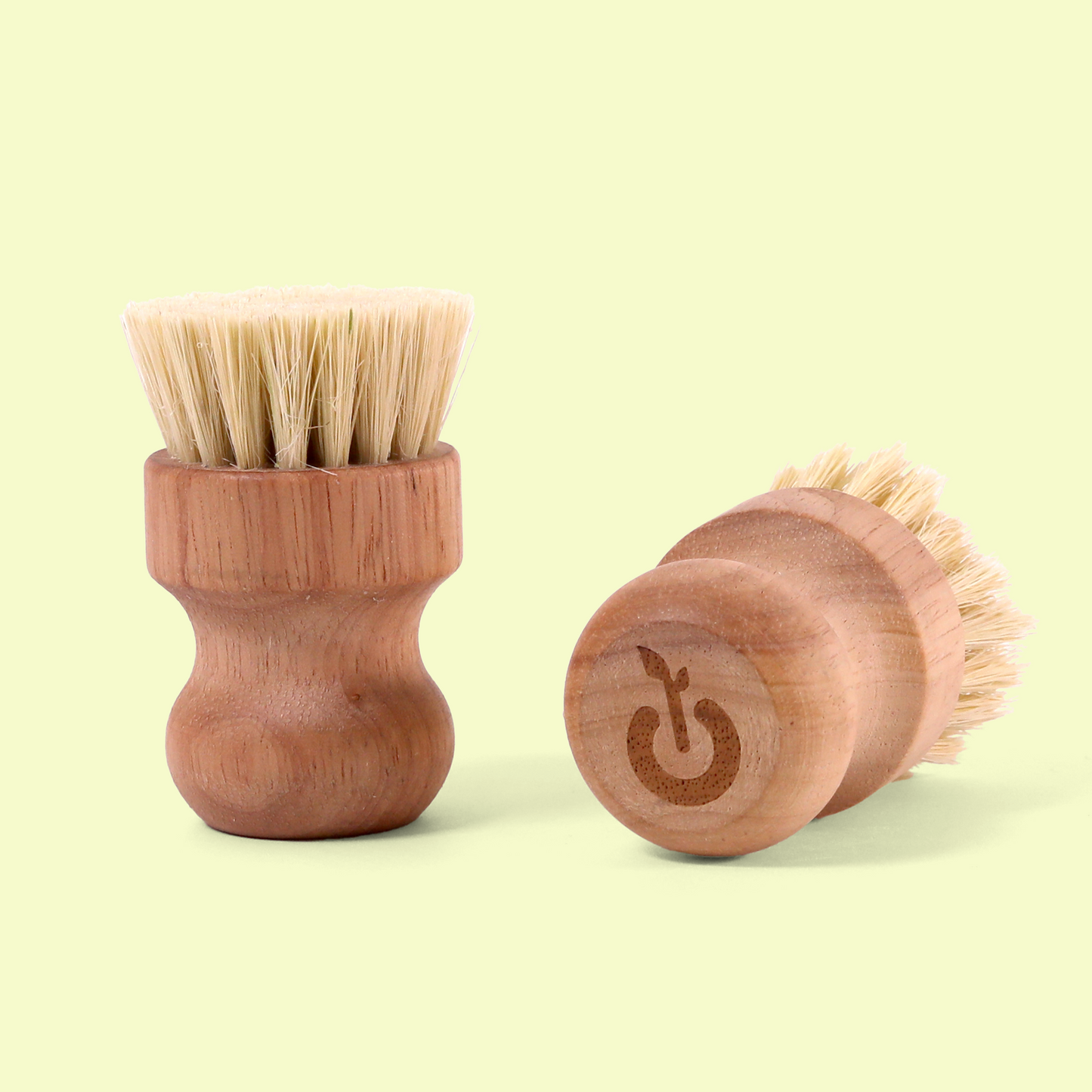 Two small wooden brushes with light-colored natural bristles. One brush is standing upright showing the bristles on top, while the other is lying on its side showing a power button/on-off symbol engraved on its base. Wood dish scrub brush from EcoGeek a Good Store product.