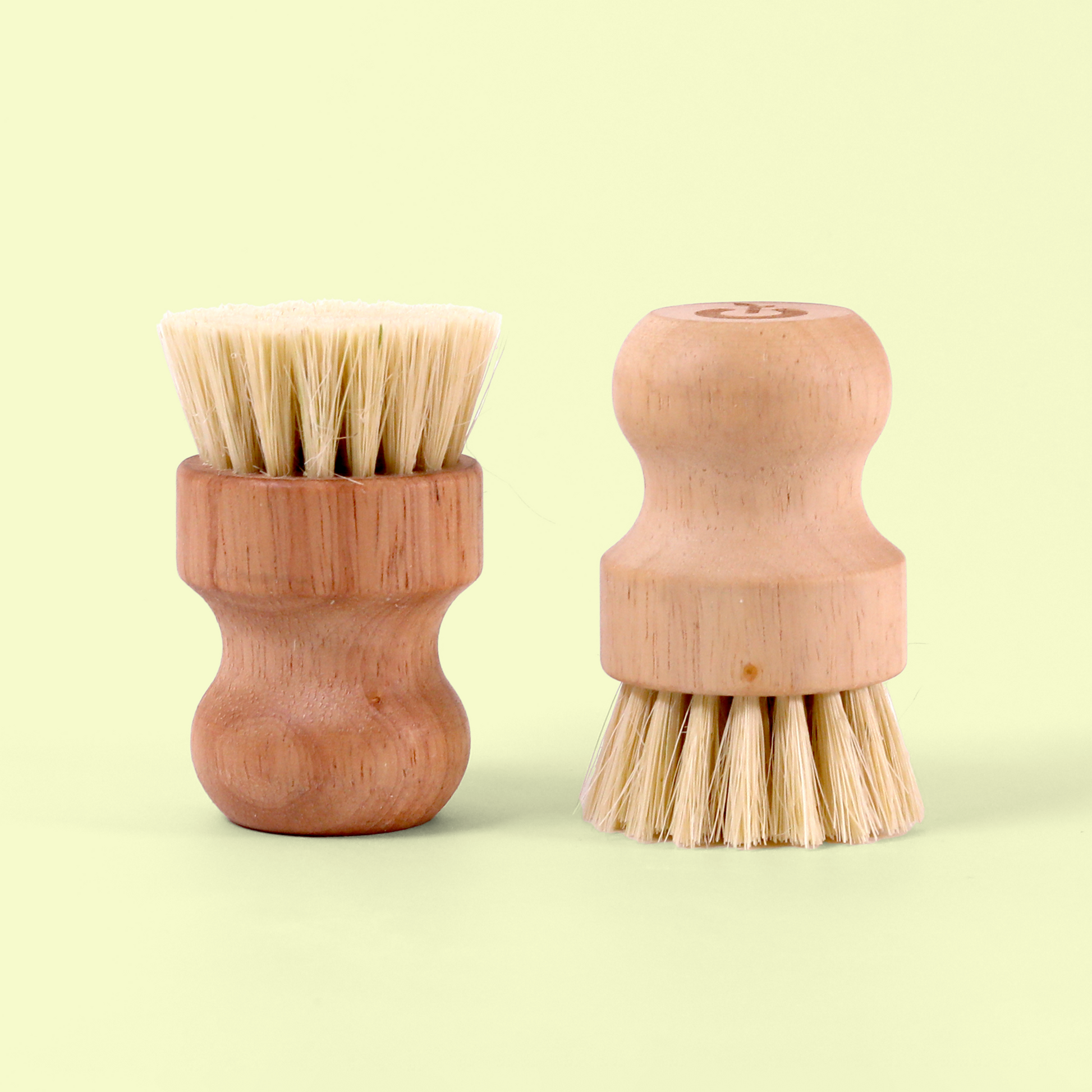 Two wooden cleaning brushes with different designs. The left brush has bristles pointing upward, while the right brush has a rounded wooden handle with bristles facing downward. Both have a similar light wood finish. Wood dish scrub brush from EcoGeek a Good Store product.