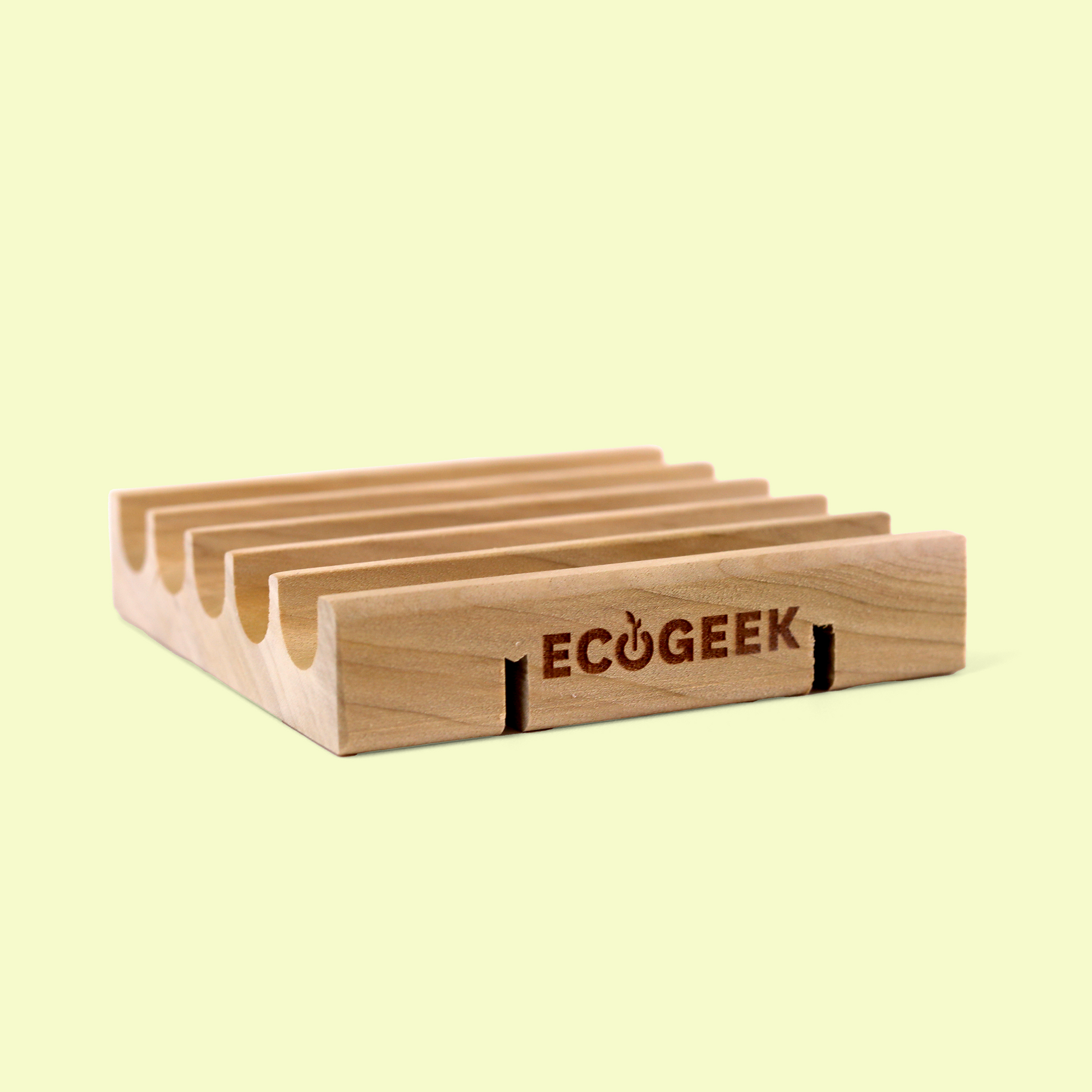 The image shows a wooden soap dish from the ECOGEEK brand displayed against a pale yellow background. The soap dish has a rectangular design with multiple parallel grooves or ridges cut into the top surface to allow water drainage and air circulation around soap bars. The soap dish appears to be made from natural, light-colored wood with visible grain patterns, emphasizing its eco-friendly design and craftsmanship. Wood soap saver from EcoGeek a Good Store product.