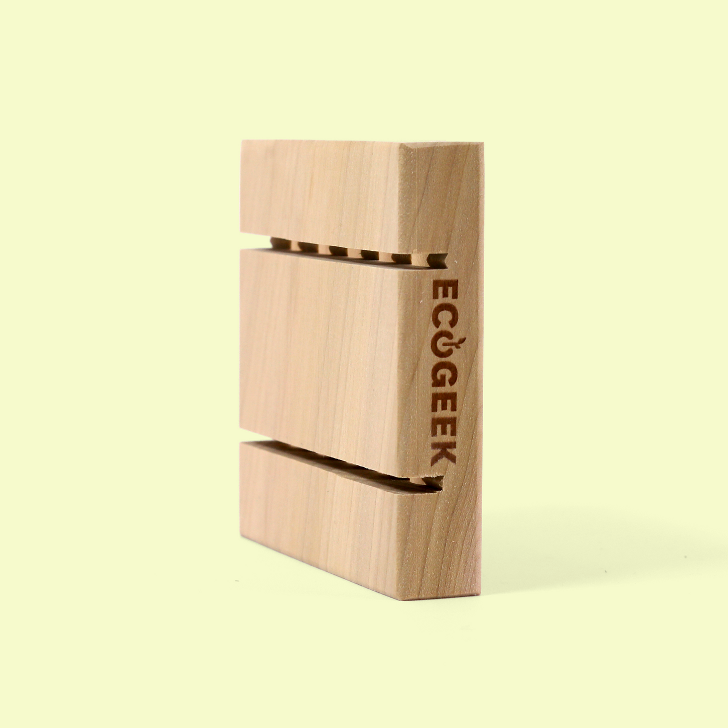 The image shows a wooden soap rack or holder from the ECOGEEK brand photographed against a pale yellow background. The holder has a vertical design with two horizontal slots cut through it, likely designed to hold a bar of soap while allowing air circulation and water drainage. The simple, minimalist design highlights the natural wood grain and emphasizes the eco-friendly nature of the product. Wood soap saver from EcoGeek a Good Store product.