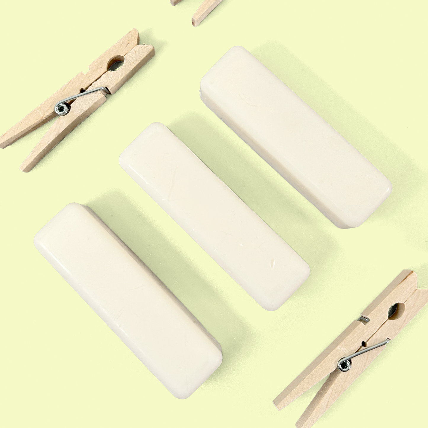 Three white rectangular soap sticks arranged horizontally in the center of the image with wooden clothespins at the corners against a pale yellow background. The soap sticks appear to be solid stain removers, with their smooth, elongated shape designed for targeted application on fabric stains. Soap stick laundry stain remover from EcoGeek and Good Store product.