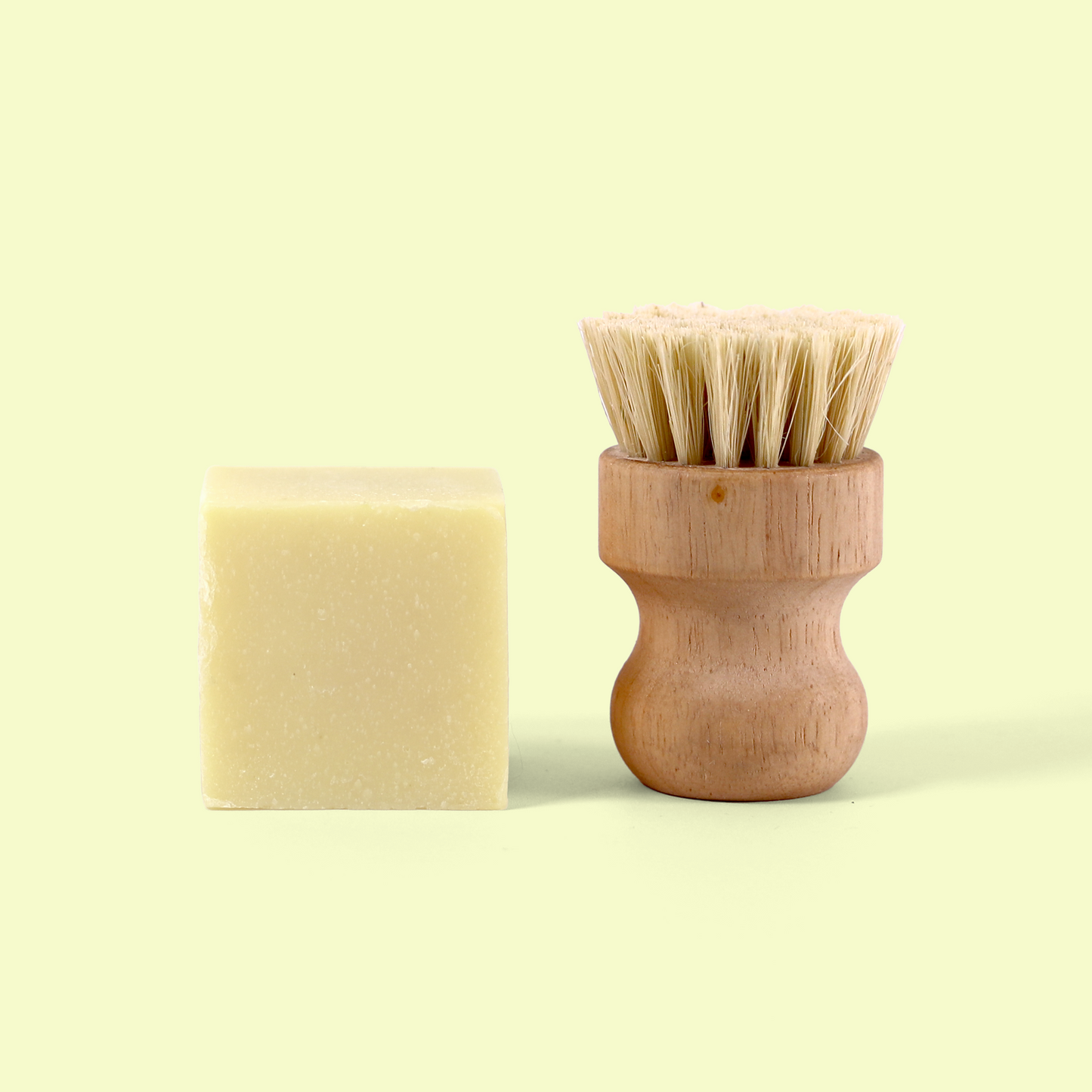 The image shows a block of cream-colored solid soap positioned next to a wooden scrub brush. The wooden brush has a curved, ergonomic handle with a round base and natural bristles protruding from the top. Both items are photographed against a pale yellow background, creating a clean, minimalist aesthetic that highlights these eco-friendly kitchen cleaning tools. Solid dish soap from EcoGeek a Good Store product.