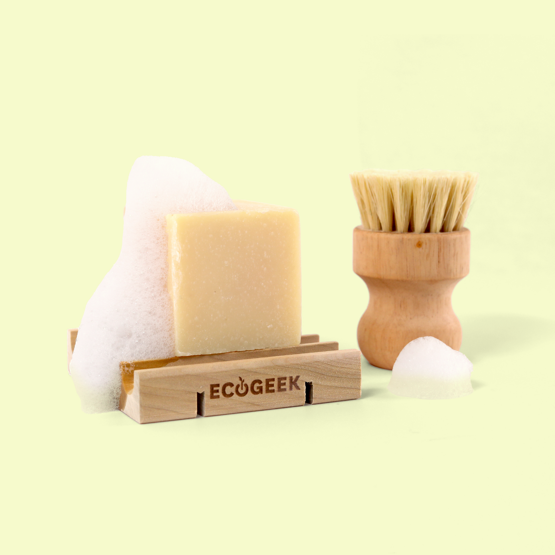 The image shows an eco-friendly dishwashing set against a pale yellow background. On the left, a cream-colored bar of solid soap sits on a wooden soap dish that has "ECOGEEK" engraved on the front. Behind the soap is a white foam or sponge. On the right is a wooden dish brush with natural bristles and an ergonomic handle. The arrangement showcases sustainable, plastic-free kitchen cleaning products from the ECOGEEK brand. Solid dish soap from EcoGeek a Good Store product.