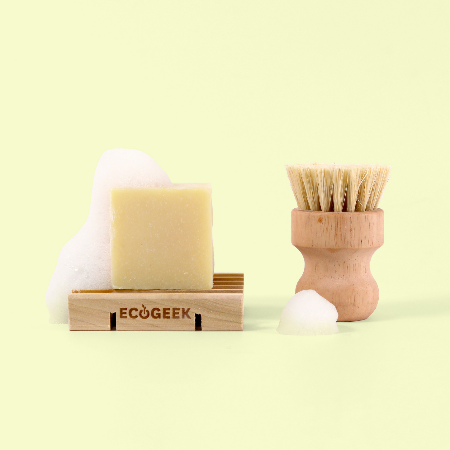 The image displays an eco-friendly dishwashing set against a pale yellow background. On the left, a cream-colored bar of solid soap sits on a wooden dish with the ECOGEEK logo engraved on the front. On the right is a wooden dish brush with natural bristles and an ergonomic curved handle.  The arrangement showcases sustainable, plastic-free kitchen cleaning products with natural materials and minimalist design. Wood soap saver from EcoGeek a Good Store product.