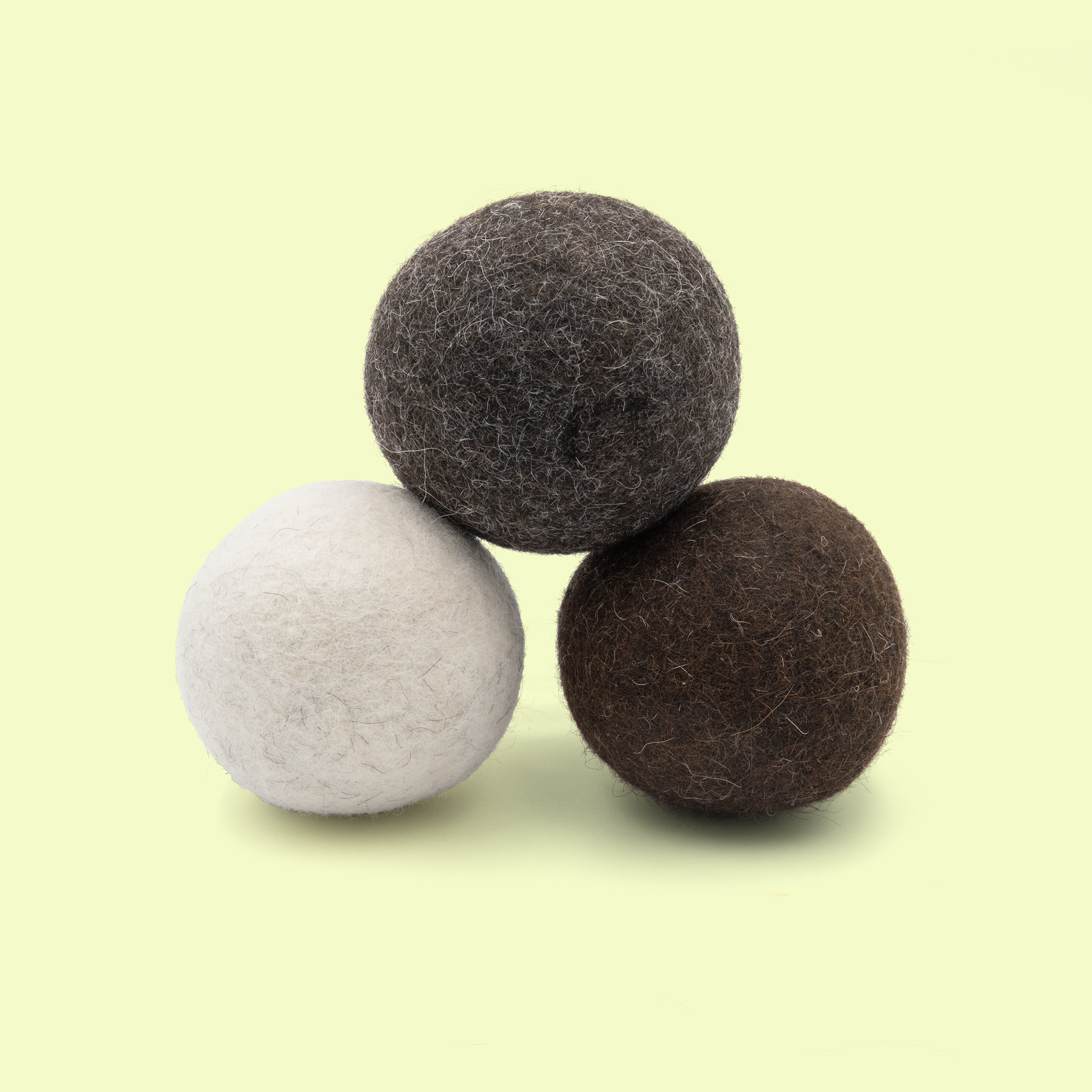Three alpaca wool dryer balls stacked in a triangular arrangement on a pale yellow background. The balls include one light gray/white ball on the left, one dark charcoal gray ball on top, and one medium brown ball on the right. All have a natural felted wool texture with visible fibers.