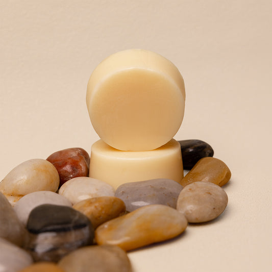Two round bars of cream-colored conditioner bars stacked on top of each other, surrounded by smooth decorative pebbles on a warm beige background.